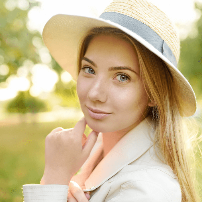 Frau mit Hut im Freien, sanft lächelnd, mit glatter Haut und natürlichem Look.