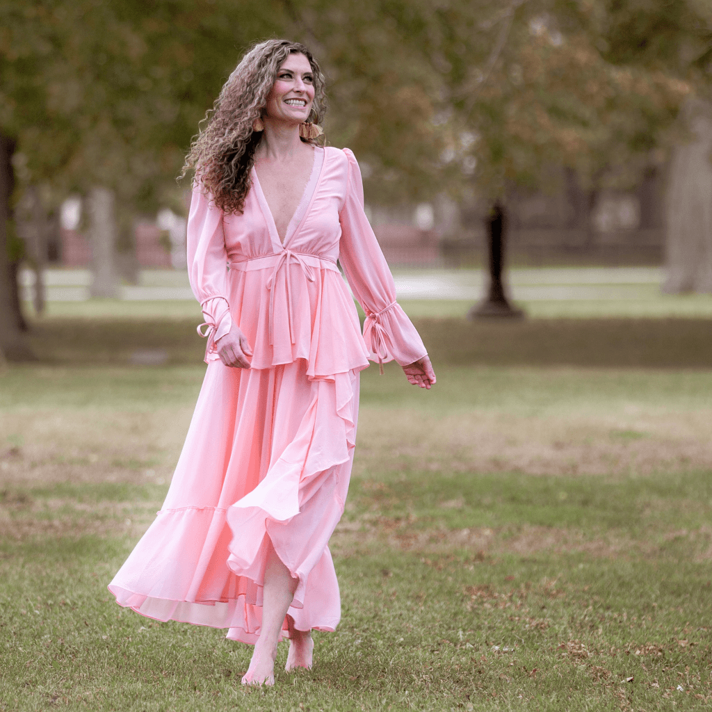 Frau in einem langen, rosa Kleid, das fröhlich durch eine grüne Wiese läuft.