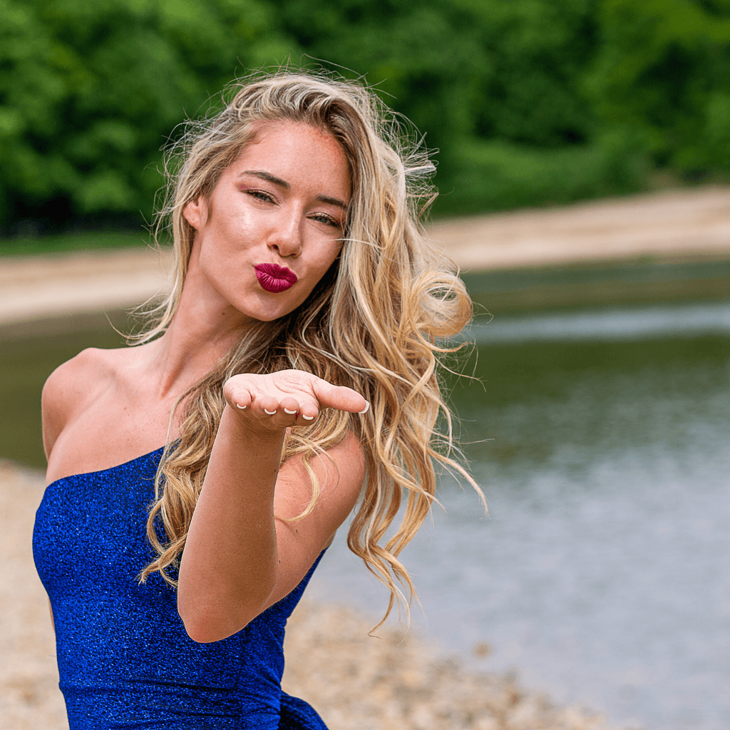 Junge Frau in blauem Kleid posiert am Wasser und bläst einen Kuss, umgeben von natürlicher Schönheit.