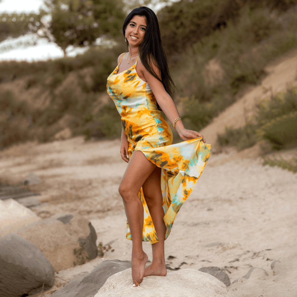 Frau in einem bunten, leichten Kleid mit einem langen Schlitz, die fröhlich auf einem Stein am Strand steht.